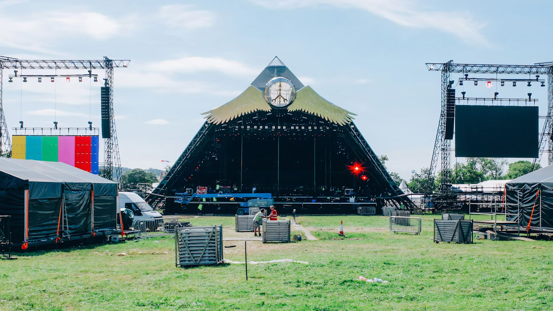 Pyramid stage at an outdoor music festival with clear blue sky and grassy field, featuring colorful lighting rigs and large screens