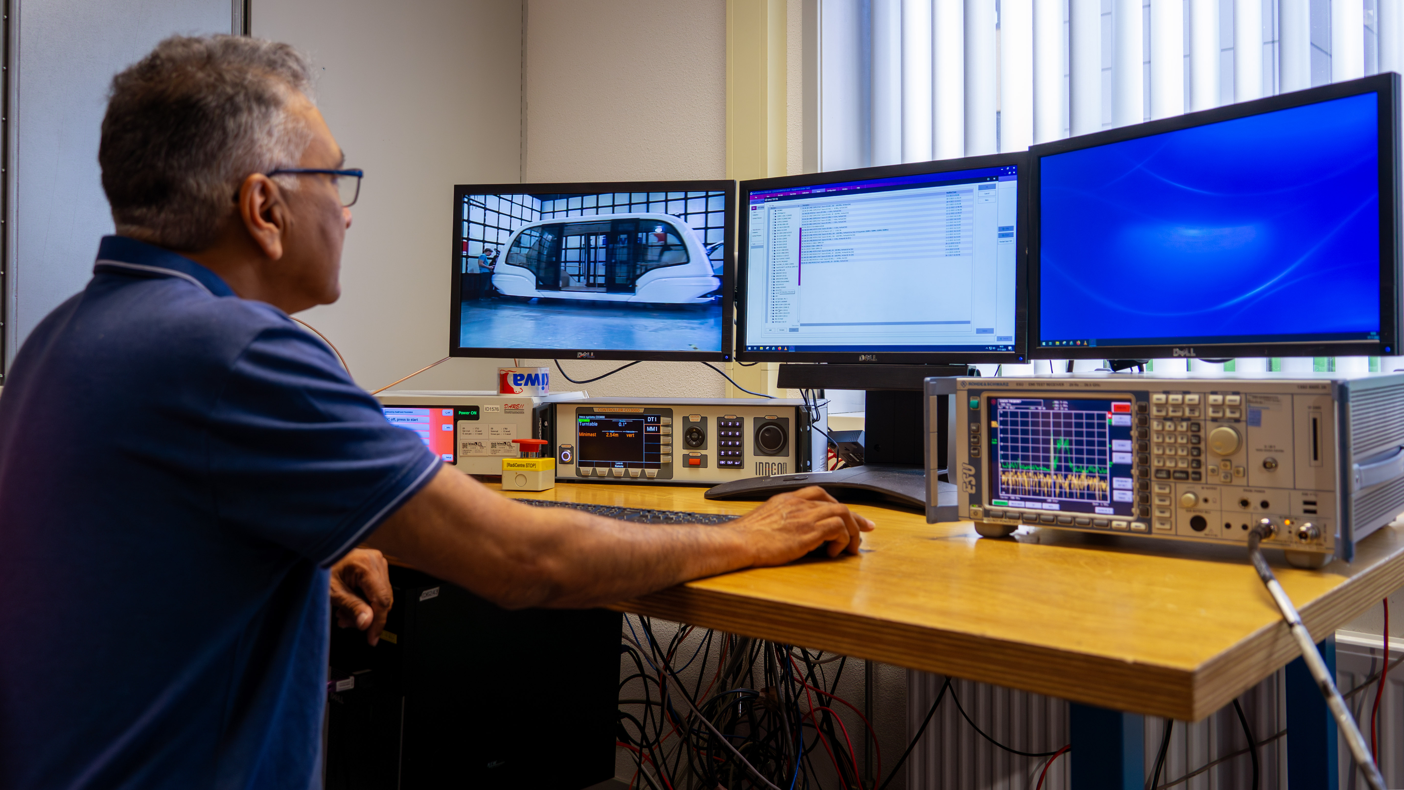 male Kiwa engineer in a technology lab working with multiple computer monitors and electronic equipment on a wooden desk, analyzing data and images of a vehicle