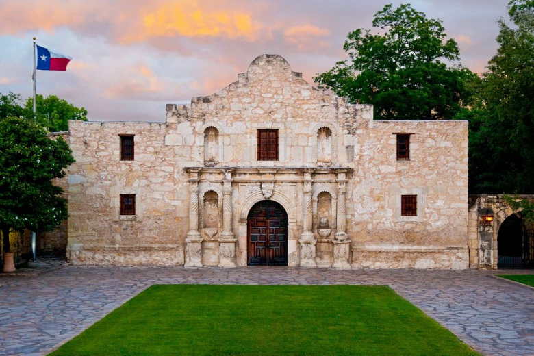 Front view of the historic Alamo Mission in San Antonio, Texas, with a Texas flag waving on the left and a vibrant sunrise sky