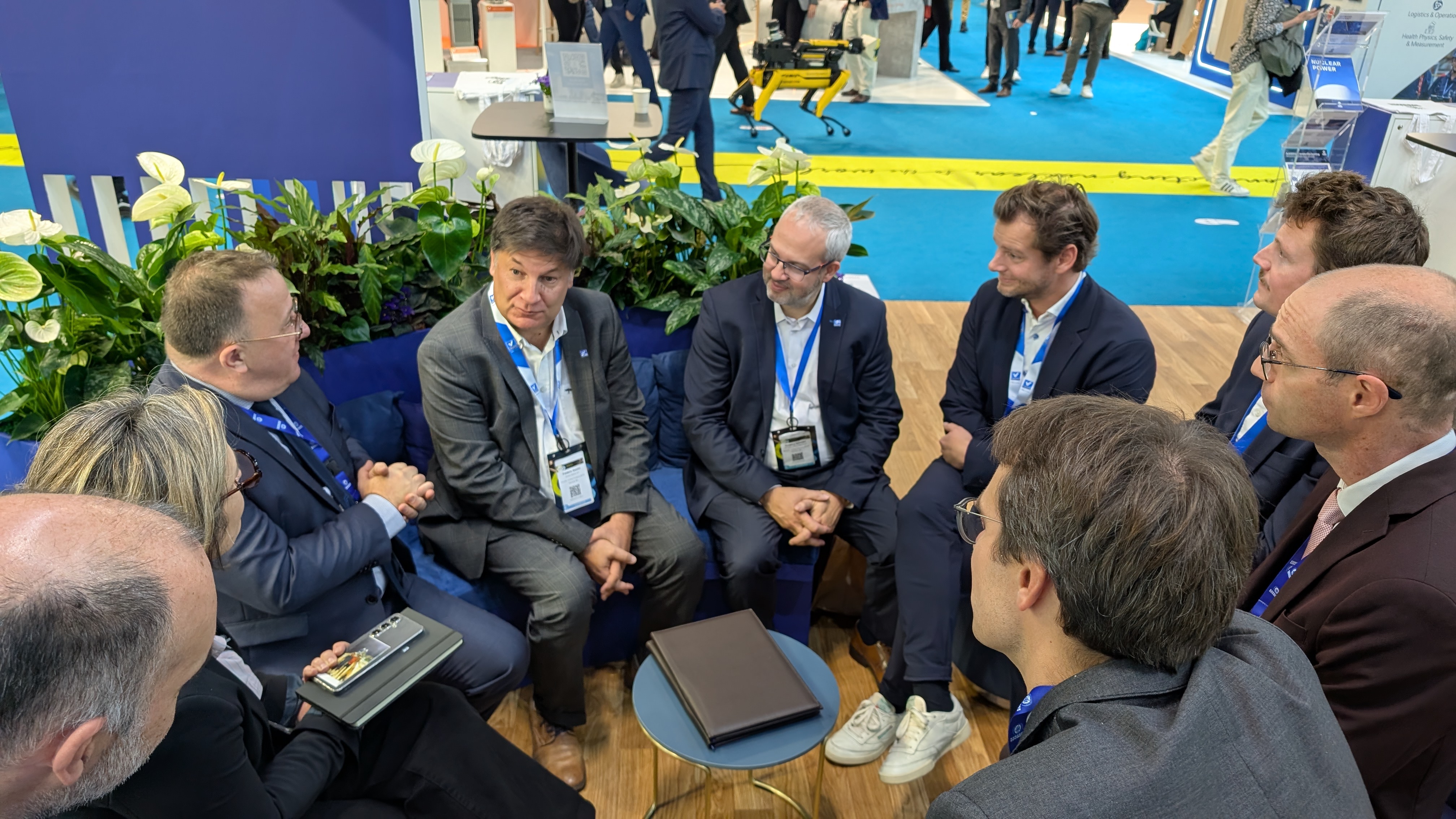 A group of business professionals in suits having a discussion at a trade show booth, surrounded by plants and a blue partition, with an exhibition floor in the background