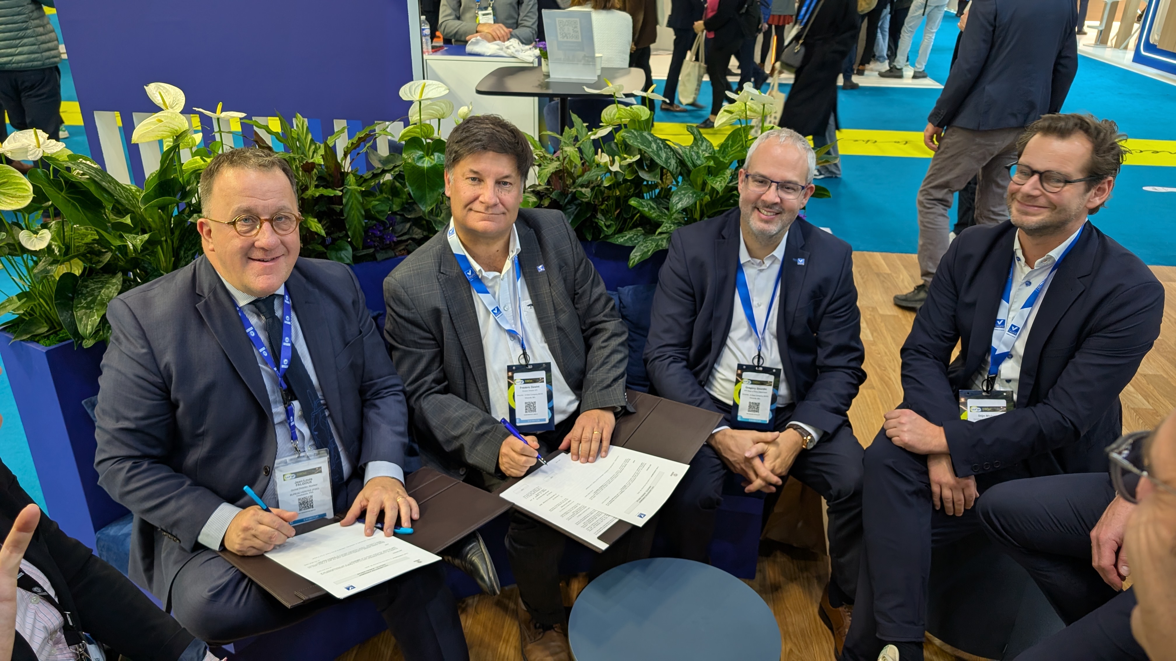 Four men in business suits seated at a table, smiling and holding documents, surrounded by greenery and a busy conference setting