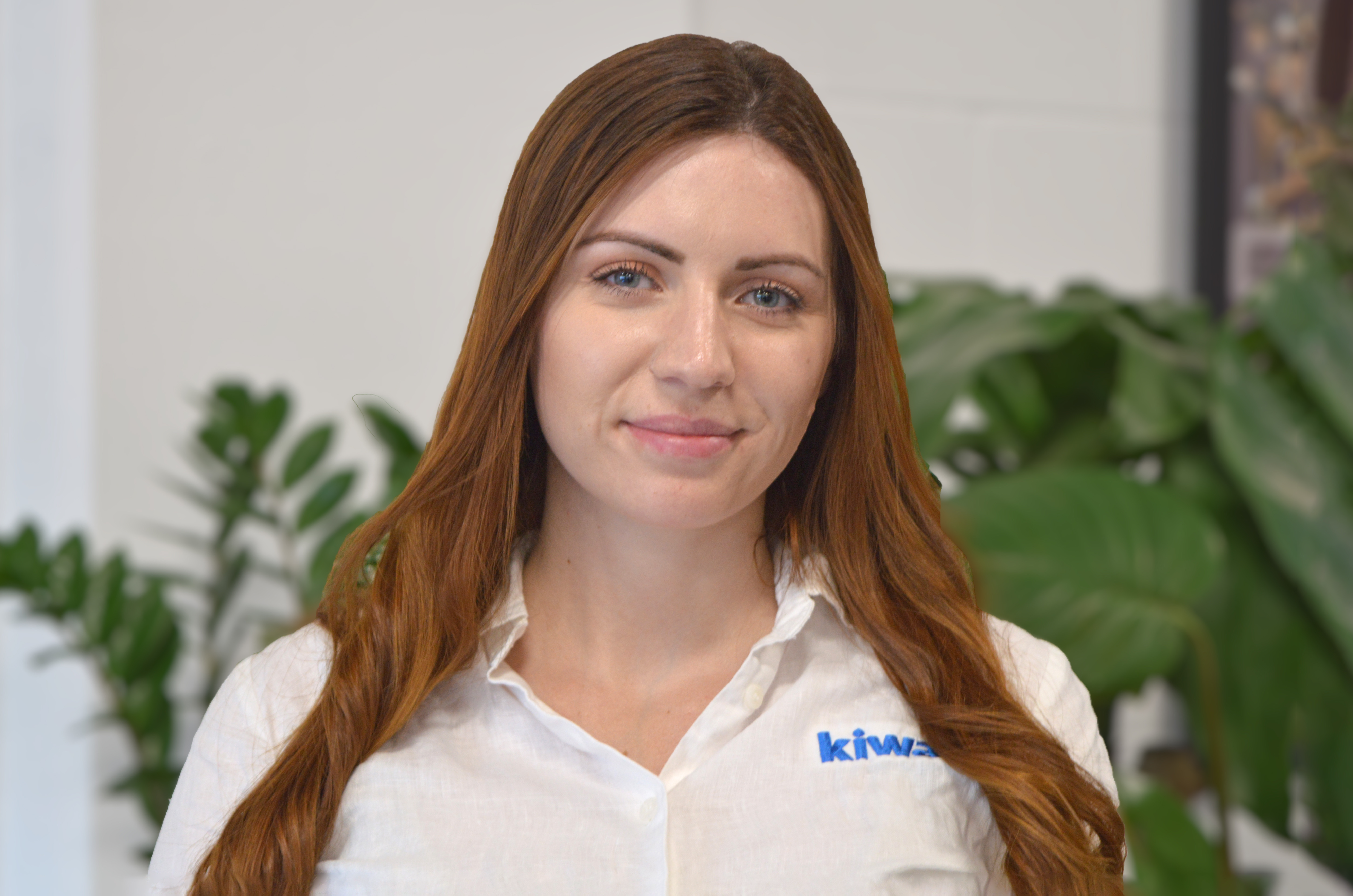 Female Kiwa professional with long red hair wearing a white shirt featuring a "kiwa" logo, standing in front of a blurred plant background