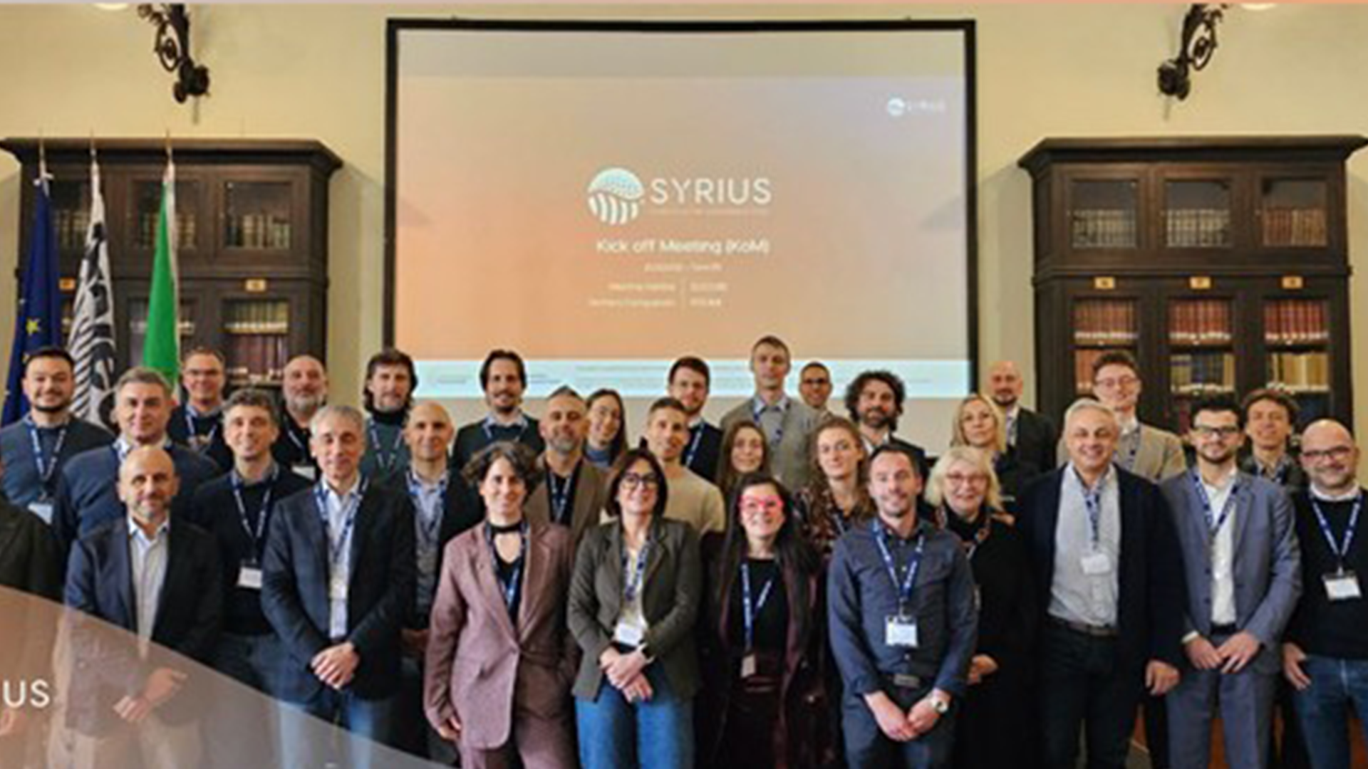 Group photo at the SYRIUS Kick Off Meeting, featuring diverse participants standing in front of a projector screen, with flags and bookshelves in the background