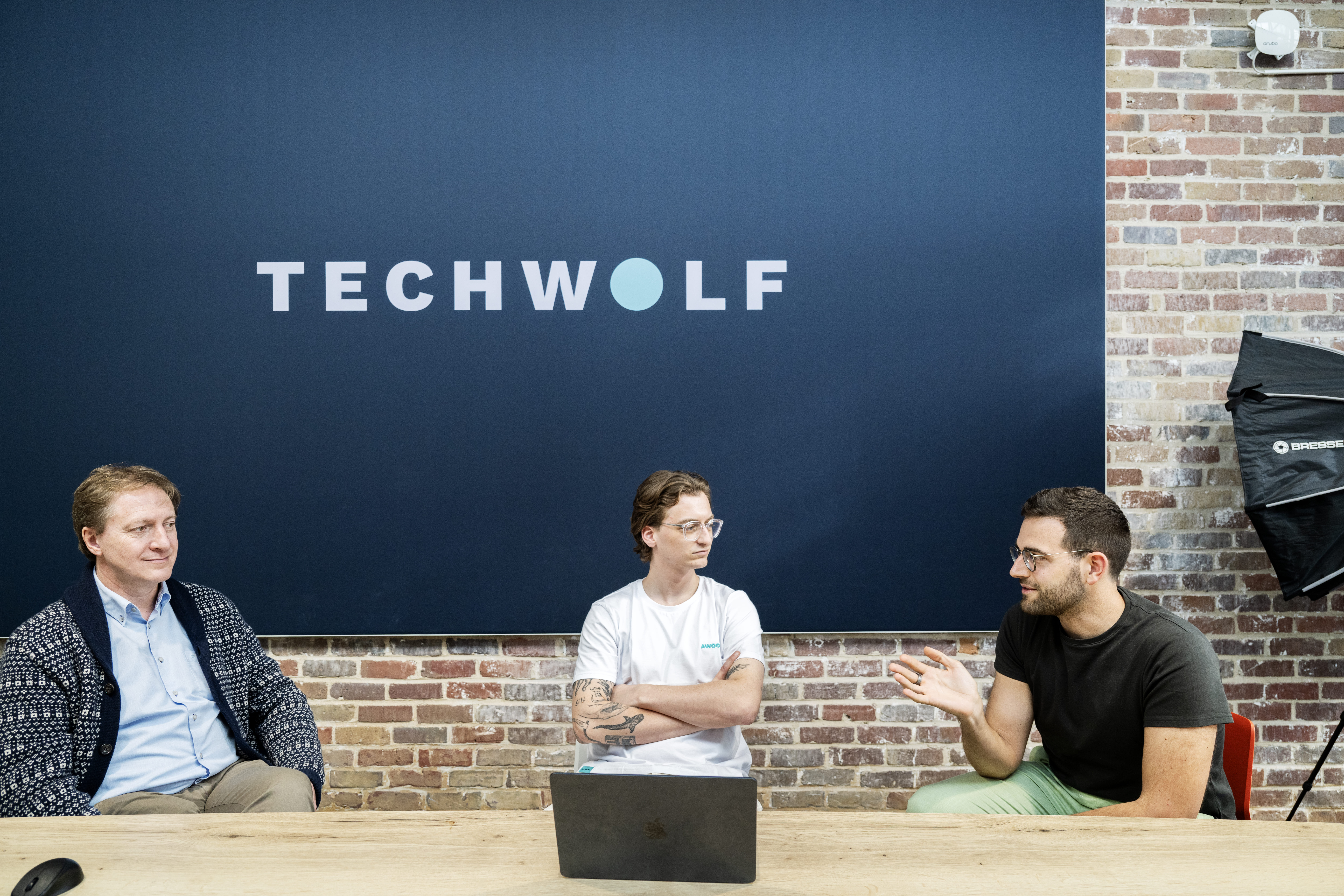 Three people engaged in discussion at a conference table, sitting beneath a large TechWolf sign on a brick wall