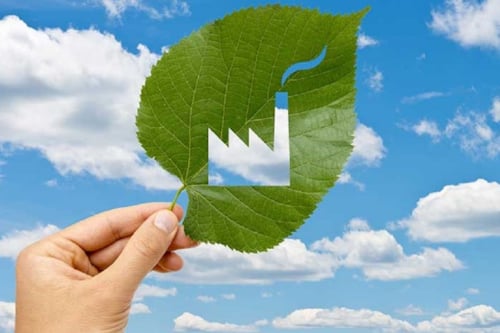 Hand holding a green leaf with a factory symbol cut out, set against a blue sky with white clouds, symbolizing eco-friendly industry practices