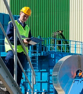 Kiwa auditor in safety gear performing an inspection with a clipboard at a manufacturing facility, featuring machinery and industrial equipment in the background