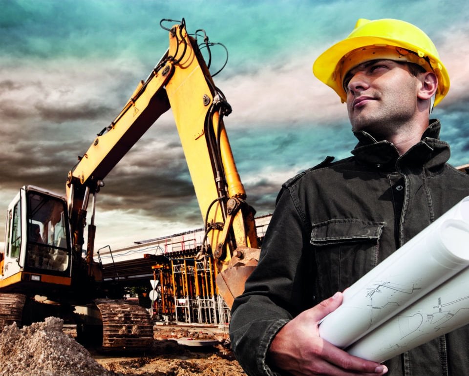 Construction site with excavator and confident worker in hard hat and holding blueprints, cloudy sky background