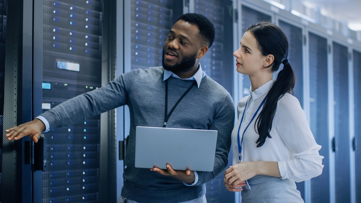 Two IT professionals in a data center discussing server maintenance while holding a laptop, surrounded by server racks