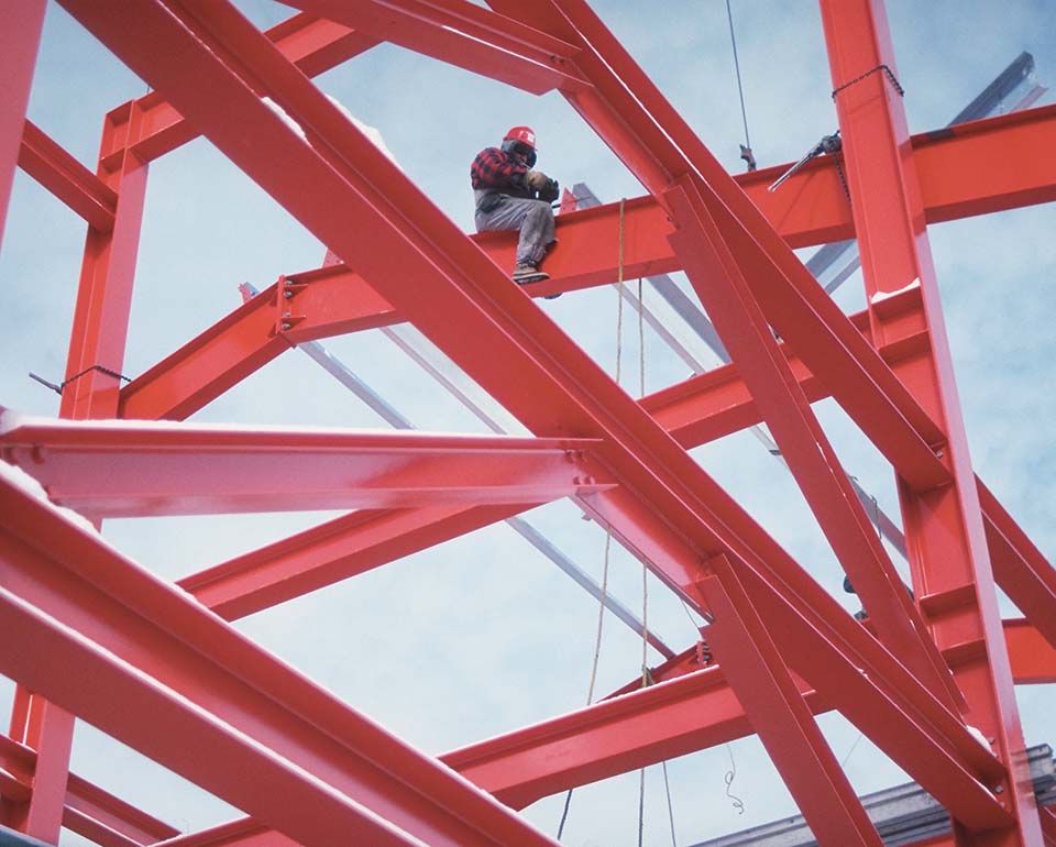 Byggnadsarbetare med skyddsutrustning sitter på en stålbalk i röd byggnadsställning mot blå himmel.