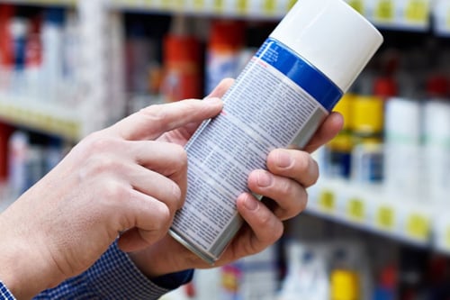 Närbild på händer när en person håller i en sprayflaska och för ett finger längst den finstilta texten på flaskans baksida för att lättare kunna läsa vad som står.