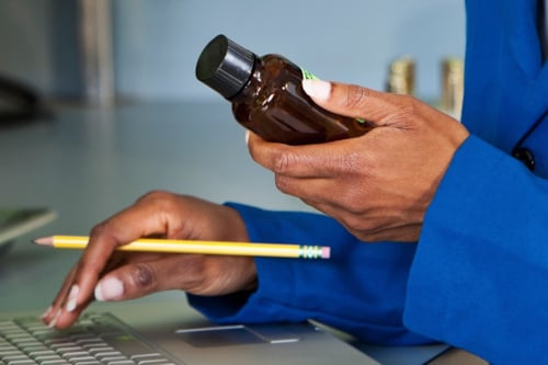 Händer syns där en kvinna håller i en liten flaska med ena handen, medan den andra handen skriver något på en laptop medan den håller i en blyertspenna mellan fingrarna.