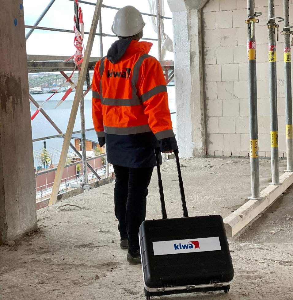 Kiwa engineer in high-visibility jacket and hard hat walking on a construction site with a Kiwa-branded suitcase, near scaffolding and a view of the water