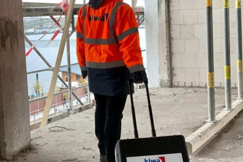Kiwa engineer in high-visibility jacket and hard hat walking on a construction site with a Kiwa-branded suitcase, near scaffolding and a view of the water