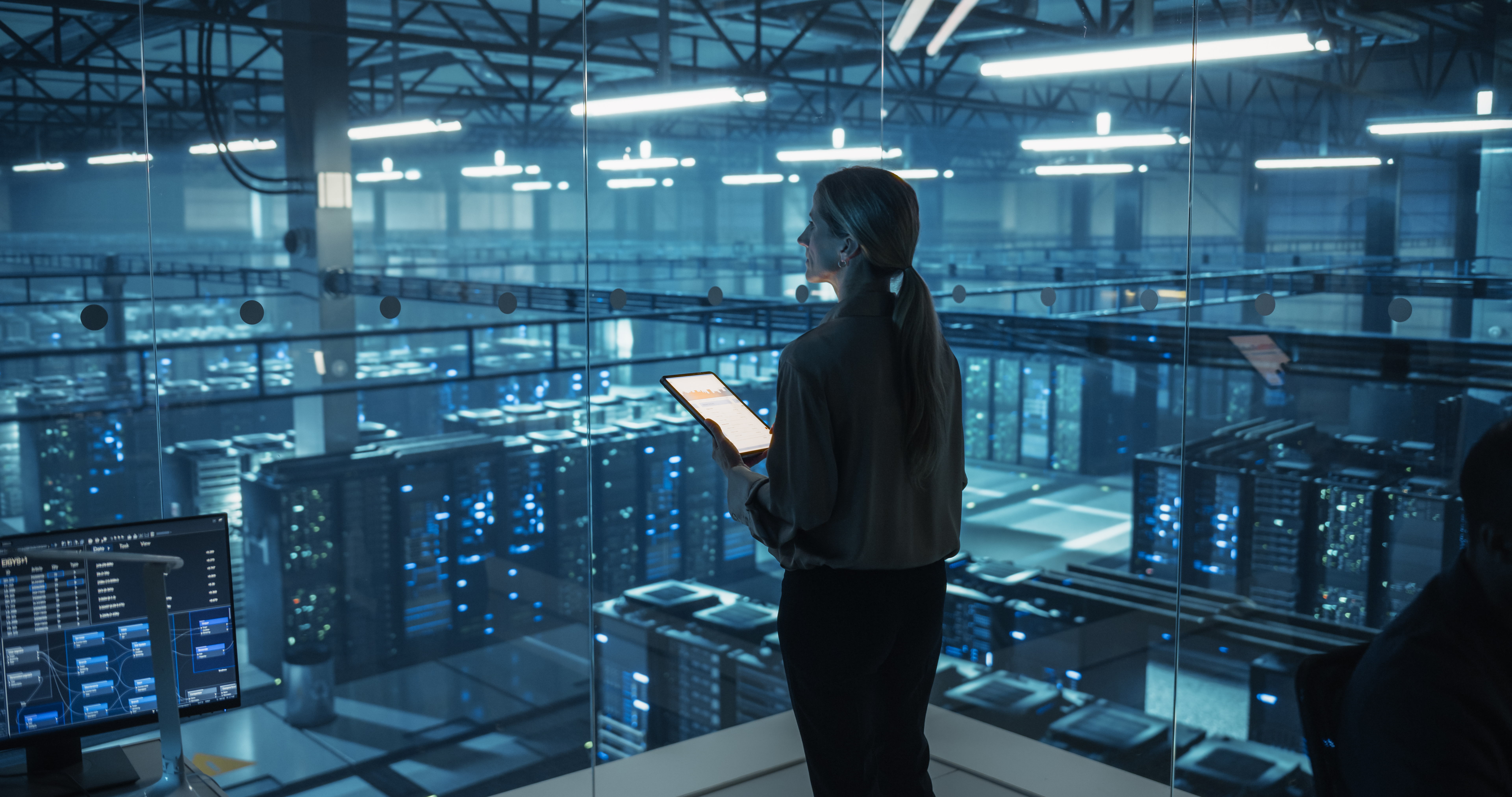 Woman using tablet in futuristic data center, surrounded by glowing server racks, monitoring digital infrastructure