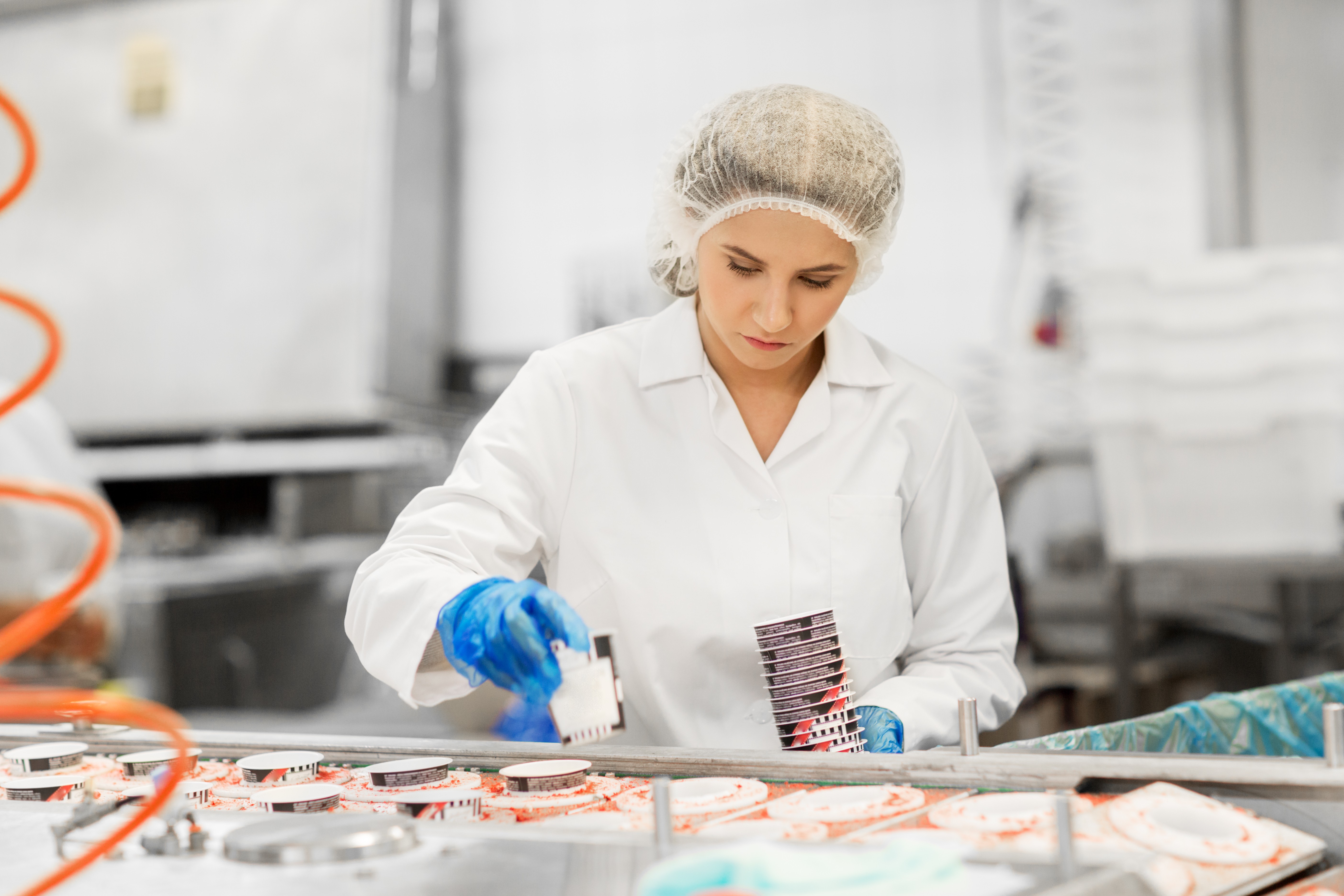 female Kiwa inspector in a white lab coat and hairnet working at a food production line, inspecting packaged products with a focused expression in a factory setting
