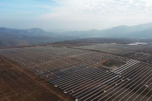 Parque solar en una región montañosa, con filas de paneles solares extendiéndose sobre un paisaje árido bajo un cielo parcialmente nublado.