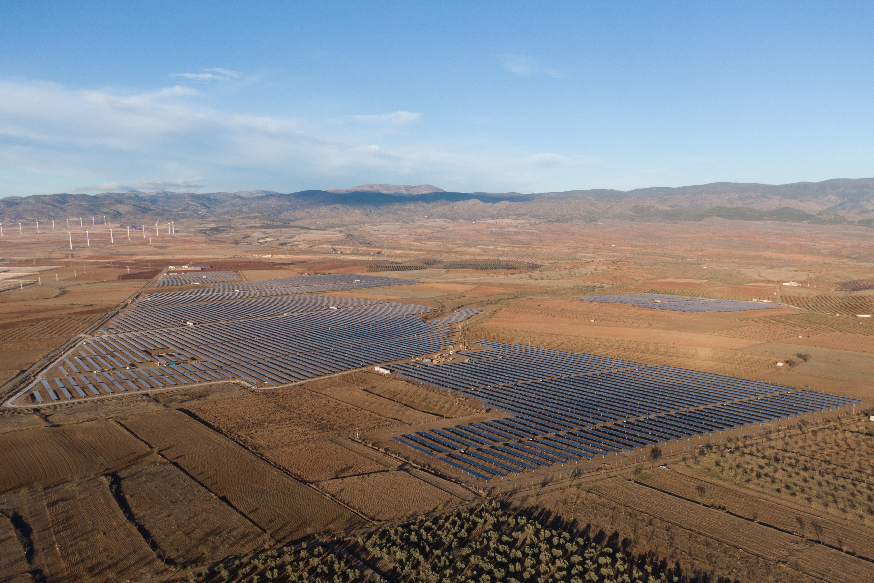 Aerial view of a vast solar farm in a rural landscape with wind turbines in the background, under a clear blue sky