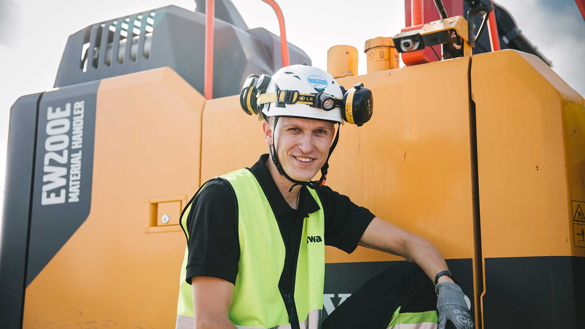 Kiwa inspektör med hjälm och gul varselväst står framför gula grävmaskiner.