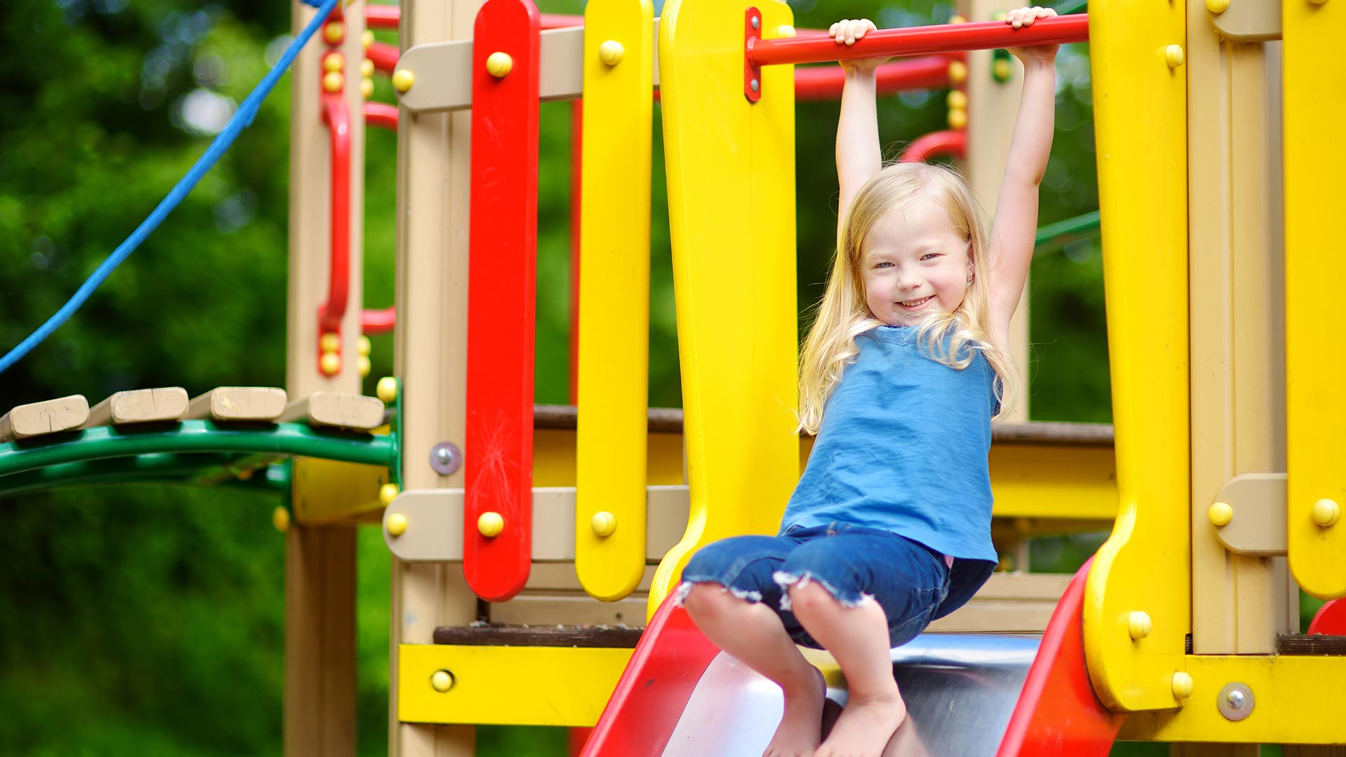 En liten flicka har roligt på en lekplats, hon håller båda händerna i en stång ovanför hennes huvud, hänger lite i den, och står barfota på rutschkanan som hon verkar vilja åka i.  