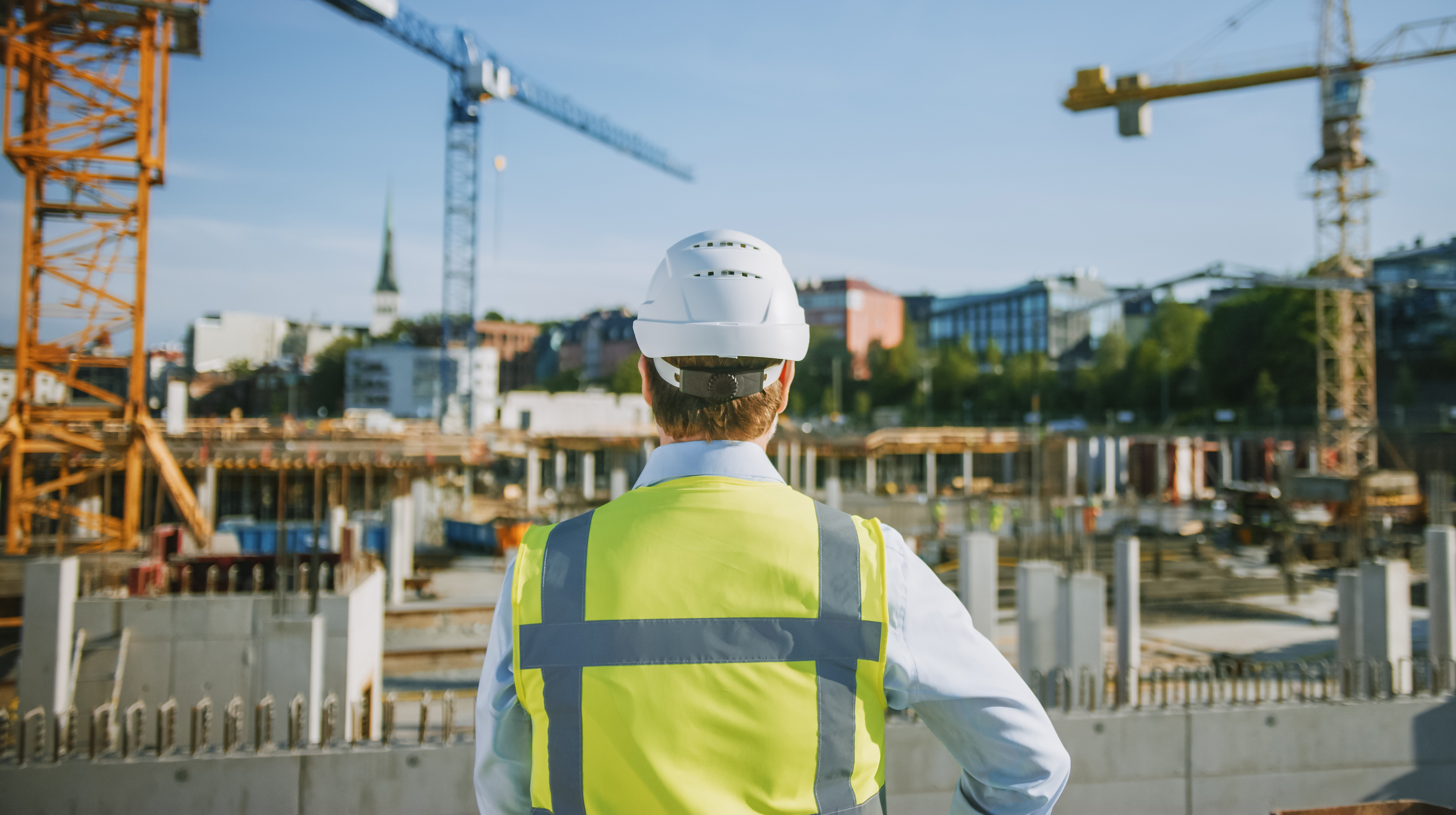En man med skyddshjälm och gul varselväst betraktar ett bygge. 