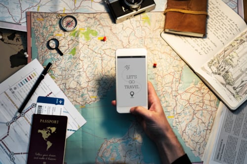 Travel planning concept with a person holding a smartphone displaying "Let's Go Travel" over a world map. Surrounding items include a vintage camera, compass, passport, and travel documents, enhancing the theme of adventure and exploration