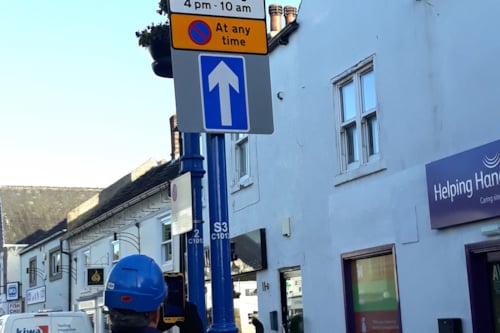 Kiwa employee inspecting street sign in urban area with one-way traffic and parking restrictions, featuring nearby buildings and service van