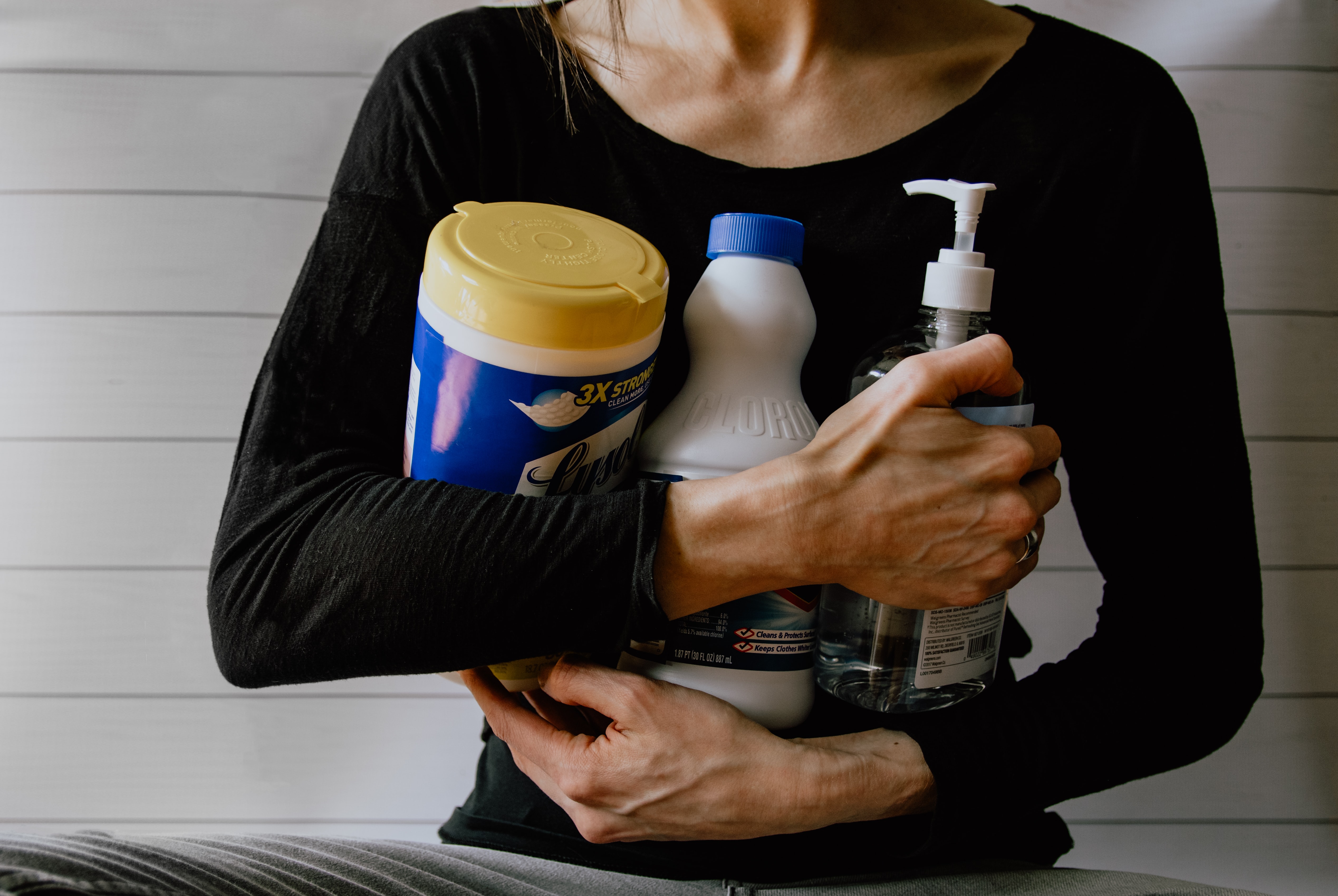 Person holding cleaning supplies including disinfectant wipes, bleach bottle, and hand sanitizer against a white paneled wall background