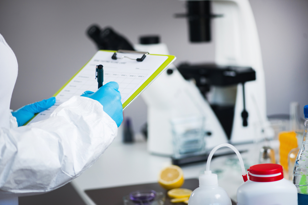 Scientist in lab coat and gloves recording data on clipboard in laboratory with microscope and equipment