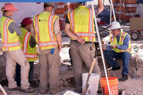 Archaeologists in safety vests and hard hats excavating a site, examining artifacts, and discussing findings under a tent