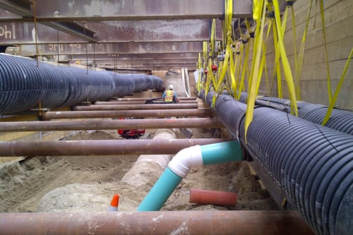 Under-construction underground utility installation site with large black pipes suspended by yellow straps, intersecting blue pipe, and construction equipment in the background