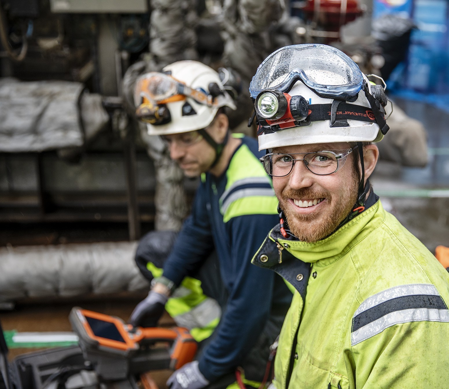 Two Kiwa inspector on-site weaaring safety gear, including helmets and protective glasses, in a factory setting. The focus is on the smiling worker in a yellow high-visibility jacket.